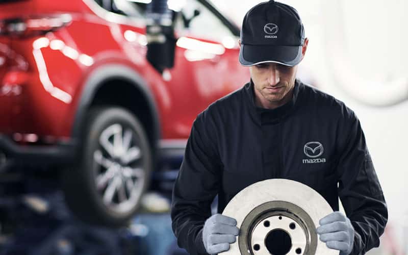 Couple and Mazda Service Tech are inspecting front tire of vehicle outside the dealership. 