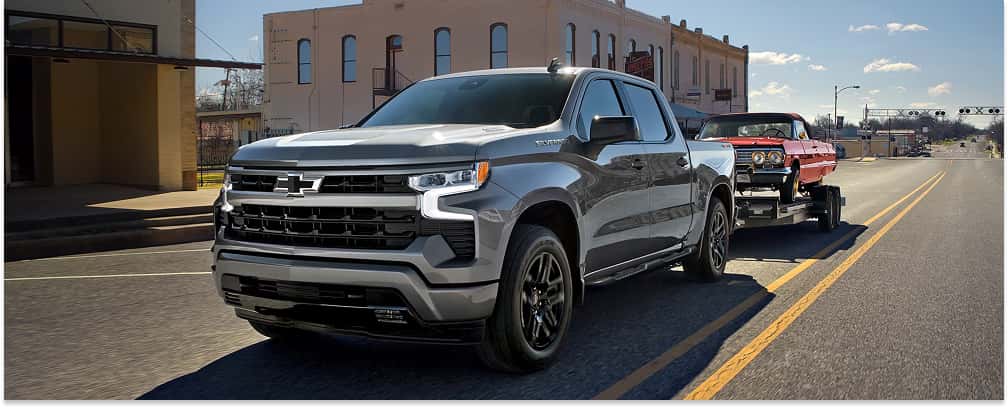 Front three-quarter view of a grey 2026 Chevrolet Silverado 1500 towing a red car trailer down a city street.