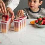 Close-up of woman and son taking out ice pop from moulds in the kitchen. Mother and son preparing mis fruit ice cream on stick. Strawberries and oranges on table.