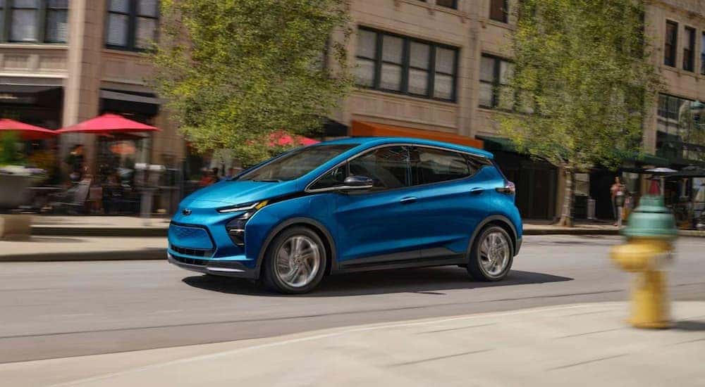 A blue 2023 Chevy Bolt EV driving on a city street past red cafe umbrellas