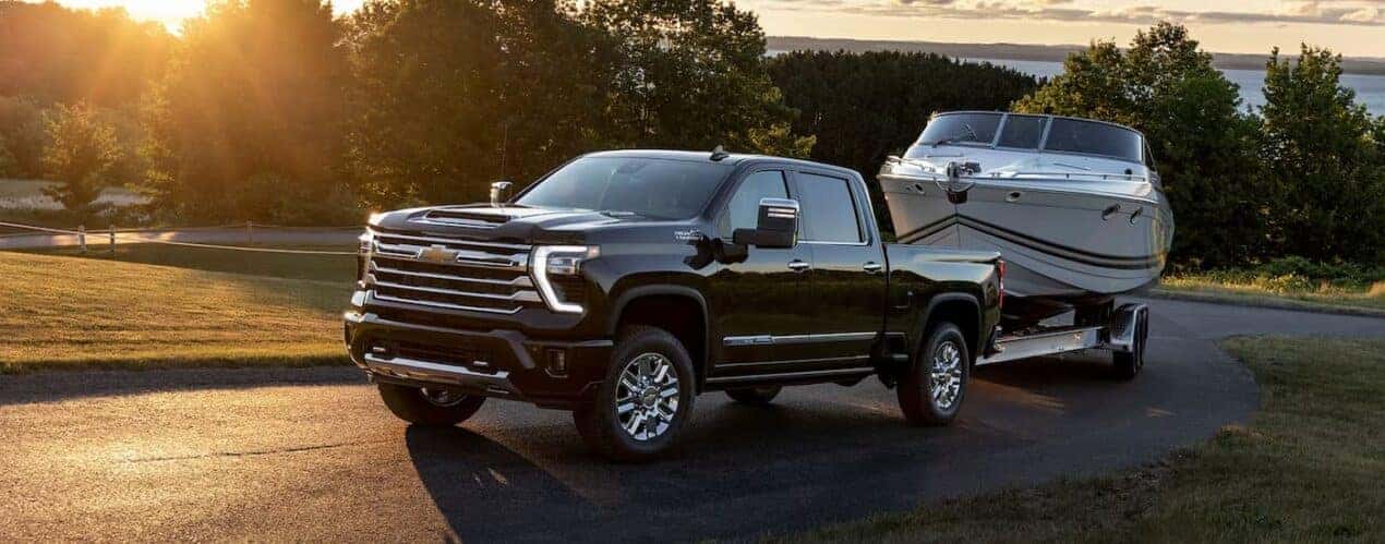 A black 2024 Chevy Silverado 2500 HD High Country is shown towing a small white boat on a winding road.