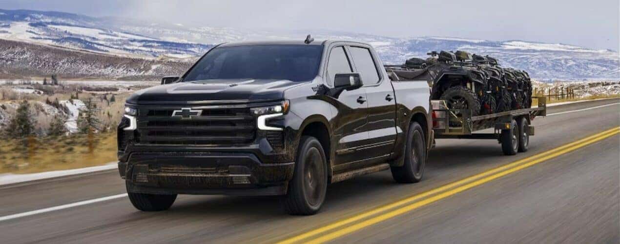 A black 2024 Chevy Silverado 1500 is shown towing a trailer on a highway.