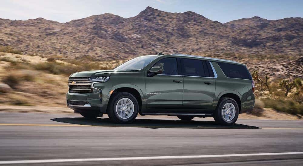 Side view of a sage green 2024 Chevy Suburban LT driving on an open road.