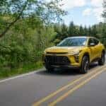 A yellow 2024 Chevy Trax ACTIV, one of many Chevys for sale near Bowling Green, driving on a tree-lined road.