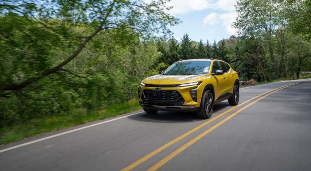 A yellow 2024 Chevy Trax ACTIV, one of many Chevys for sale near Bowling Green, driving on a tree-lined road.