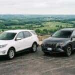 A realistic photograph of two modern compact crossover SUVs parked side by side on a paved overlook, one a white Chevrolet Equinox and one a dark grey Hyundai Tucson, three-quarter front angle showing both vehicles clearly, rolling green Kentucky countryside and an overcast sky in the background, even natural daylight. No text, no logos other than natural vehicle badging, no identifiable faces. (Representative image for illustration purposes only. Actual vehicle may vary based on trim and configuration.)