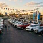 A realistic wide photograph of a Chevrolet dealership lot at golden hour in a small Kentucky town, rows of new Chevrolet pickup trucks and SUVs parked in neat lines with clearance-season pennant flags strung overhead, a sales building with glass showroom windows in the background, warm late-afternoon light, shallow depth of field. No text, no logos other than natural vehicle badging, no identifiable faces. (Representative image for illustration purposes only. Actual vehicle may vary based on trim and configuration.)