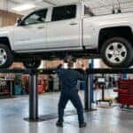 A realistic photograph inside a clean, well-lit automotive service bay, a modern silver full-size Chevrolet pickup truck raised on a hydraulic lift, a technician in plain work coveralls seen from behind inspecting the four-wheel-drive transfer case and driveshaft underneath, tools and diagnostic equipment nearby, bright overhead shop lighting. No text, no logos other than natural vehicle badging, no identifiable faces. (Representative image for illustration purposes only. Actual vehicle may vary based on trim and configuration.)