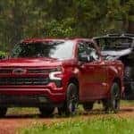 A red 2025 Chevy Silverado 1500 RST is shown towing a trailer off-road.