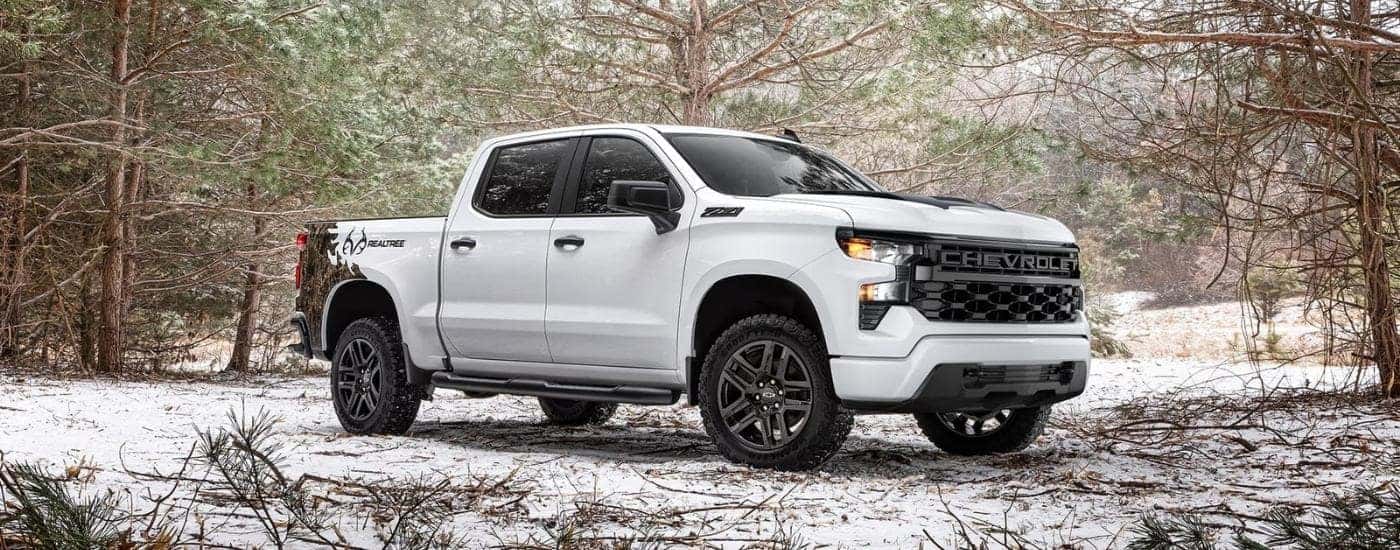 A white 2024 Chevy Silverado 1500 Realtree Edition is shown parked in a snowy clearing.