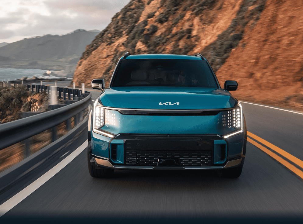 A blue 2025 Kia EV9 driving on a coastal highway at sunrise